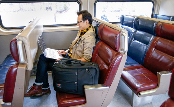 A person using a laptop while sitting in a rail car seat with a Black 525 Ballistic Western Flyer in the seat beside them.